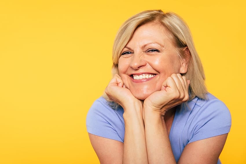 Mature woman stands in front of a yellow background, showing her amazing non-surgical facelift off